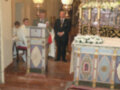 En una iglesia, un sacerdote se prepara para oficiar la misa. En el altar, flores blancas adornan un manto de color azul y dorado. Detrás del altar, una imagen religiosa se destaca en un marco dorado. A la izquierda, un hombre con una saya blanca se prepara para subir al altar. La imagen está iluminada por una lámpara de cristal y un farol, creando un ambiente reverente.