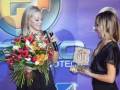 Dos mujeres en un evento, una con flores y la otra con un trofeo.