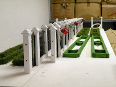 Un pequeño jardín de miniaturas con un muro blanco, plantas verdes y cajas de cartón en el fondo.