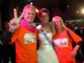 Un hombre con una camiseta naranja y un hombre con una corbata rosa dan la señal de "OK". Una mujer con un vestido blanco y una corona llamada "Miss España" sonríe junto a ellos. En el fondo, se pueden ver otras personas y luces de neón.