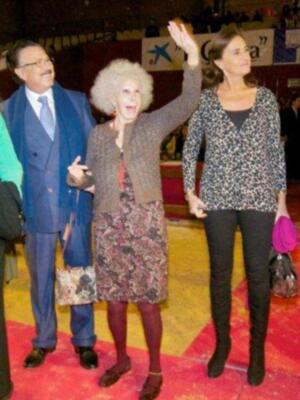 Tres personas saludan en una ceremonia o evento público en un escenario con público en el fondo.