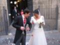 Feliz pareja recién casada saliendo de una iglesia, rodeados de confeti rojo y blanco. El novio lleva un traje negro y corbata roja, mientras la novia lleva un vestido blanco con mangas largas y un ramo de rosas blancas.