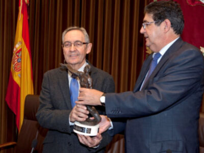 Dos hombres en trajes celebran un momento de reconocimiento, uno entrega un trofeo mientras el otro sonríe. La bandera española y una alfombra roja completan la escena formal.