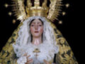 Una figura religiosa vestida con un manto dorado y una capa blanca, rodeada de estrellas doradas. La figura lleva un manto con detalles en oro y una corona brillante, sosteniendo un objeto en su mano izquierda. La figura es parte de una procesión religiosa, probablemente representando a la Virgen María.