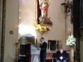 Un hombre se dirige a una audiencia en un ambiente formal, posiblemente una iglesia o sala de conferencias. La imagen muestra a un hombre en traje negro al frente, mientras otro se sienta en una silla cerca de él. La decoración incluye un altar con flores y un crucifijo en la parte superior, iluminado por una lámpara. La atmósfera es serena y respetuosa, indicando una ceremonia o discurso importante.