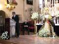 En una iglesia, un hombre en traje negro se dirige a una audiencia. A su lado, otra persona está sentada en un banco rojo. En el fondo, una imagen religiosa majestuosa se destaca entre flores blancas y velas. La arquitectura y decoración sugieren una celebración o servicio religioso significativo.