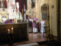 En una iglesia, un sacerdote en vestido morado y otro en blanco se encuentran frente a una mesa alta con velas encendidas y flores. A la derecha, un altar con una imagen de San Juan Evangelista. La atmósfera es solemnemente reverente, típica de una ceremonia religiosa.