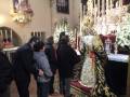 Una imagen de una iglesia con una Virgen María vestida en blanco y dorado, rodeada por fieles rezando. La decoración es elaborada con flores y velas, creando un ambiente de reverencia.
