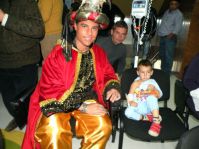 Un hombre vestido con un traje rojo y dorado, sosteniendo una figura de juguete en brazos. Al fondo, otros individuos observan la escena.