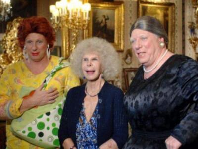 Tres personas posan en una sala decorada con luces y cuadros. La persona de la izquierda lleva un vestido amarillo con una bolsa verde y blanca, la central tiene un vestido azul con detalles dorados y la derecha una vestimenta negra.