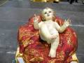 Un bebé Jesús en un vestido blanco y dorado, sentado sobre una almohada roja con bordados dorados.