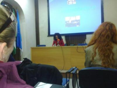 A una persona hablando en un escenario con una pantalla proyectada en el fondo. La persona está usando auriculares y tiene un micrófono en la mano izquierda. En el fondo, se puede ver una persona sentada frente a un escritorio con varios dispositivos de audio. La escena parece ser parte de una conferencia o presentación.