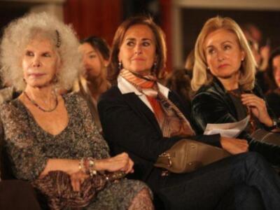 Una escena en un evento formal con varias personas sentadas, destacando a una mujer mayor en el centro con una bufanda y una mano en la cadera, mientras otras observan.
