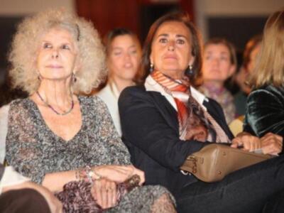 Una mujer con cabello gris y una americana de lana es la protagonista, mientras que otras mujeres observan atentamente.