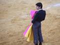 Un torero en traje tradicional de color azul, con un capote amarillo y rosa, preparándose para la corrida de toros en una arena de tierra.