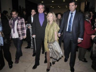 Una imagen de una celebridad caminando entre otros personajes. La celebridad lleva un traje negro, una chaqueta de piel y una bufanda. La celebridad es seguida por otros personajes, algunos con abrigos y chaquetas. La escena parece ser en un evento o fiesta formal, con luces y decoración de fondo.