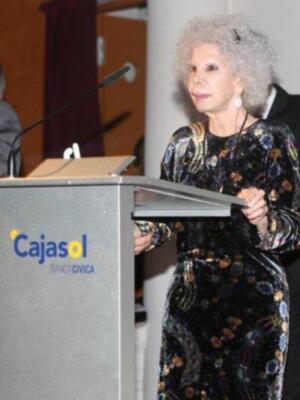 Una mujer con cabello blanco y largo, vestida con un traje floral negro, se encuentra en una conferencia o evento. Está frente a un podio con la marca "Cajasol" y el texto "BANCA CIVICA". La imagen parece ser de un evento formal, posiblemente una presentación o discurso.