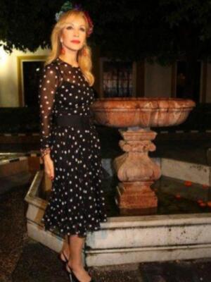Una mujer con vestido negro de lunares blancos, cabello largo y rubio, lleva un collar rojo. Detrás de ella hay una fuente con agua y sombras en el suelo.