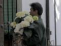 Un hombre sostiene un ramo de flores blancas en una mano, frente a una puerta con persianas verdes.