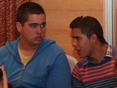 Dos jóvenes conversan en un ambiente interior, uno con una sudadera azul y el otro con una camiseta a rayas. La imagen sugiere un ambiente casual o familiar, posiblemente en una sala de estar o comedor.
