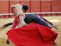 Un torero en traje tradicional lucha con un toro negro en una plaza de toros. El torero usa un capote rojo y un casco ornamental, mientras el toro lucha con él. La arena es de tierra y hay una cerca roja en el fondo, con espectadores sentados en los asientos.