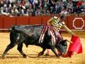 Un toro negro con una correa roja y blanco en su espalda, un matador de toros con chaqueta amarilla y negro, y una bandera roja en el fondo.