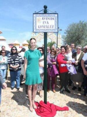 Una mujer en un vestido verde se encuentra sobre una placa de calle con el nombre "Avenida Eva González". La imagen muestra a varias personas observando la escena.