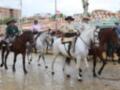 Hombres montando caballos en una carrera tradicional, con cascos y sombreros. Fondo de edificios y decoración festiva.