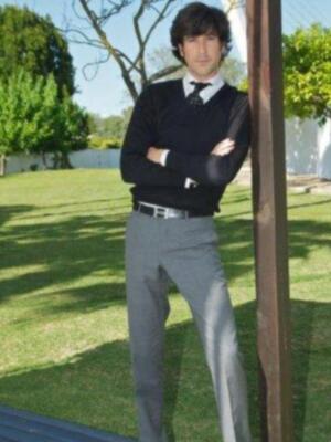 Un hombre con una camisa blanca, un suéter negro y pantalones grises posa en el jardín. La imagen muestra un ambiente tranquilo con árboles y césped verde en el fondo.