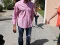 Un hombre en una camisa rosa y jeans está caminando por un camino de baldosas. En el fondo, hay una casa con plantas y otro individuo en la distancia.