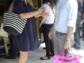 Una pareja camina por una calle, con una mujer llevando una bolsa de mano rosa y un hombre con una camisa blanca y pantalones grises.