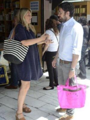Una pareja camina por una calle, con una mujer llevando una bolsa de mano rosa y un hombre con una camisa blanca y pantalones grises.