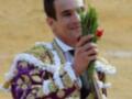 Un torero sonríe mientras sostiene una ramita de flores rojas y verdes, vestido con un traje tradicional de color púrpura y dorado. La imagen captura el momento de un torneo taurino, con una arena de arena en el fondo.