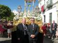 Dos hombres en trajes posan frente a una procesión religiosa con altares dorados y flores. El cielo es claro y el ambiente parece ser de una celebración festiva o religiosa importante.