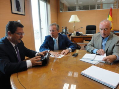 Tres hombres en trajes sentados en una mesa de oficina, discutiendo documentos y usando tabletas digitales.