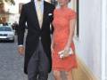 Una pareja elegante camina por una calle adoquinada. La mujer, con un vestido naranja brillante y cabello recogido, sostiene una pequeña bolsa. El hombre, con traje negro y corbata amarilla, camina junto a ella. Detalle de una celebración formal en un entorno urbano con palmeras y edificios a la vista.
