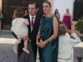 Familia formal en evento, hombre con traje y corbata, mujer en vestido verde, niña en brazos del hombre.