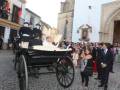 Un hombre en traje negro se sienta en un carruaje de caballo mientras una multitud observa desde el lado. La imagen muestra detalles arquitectónicos de un edificio histórico y una iglesia en el fondo.