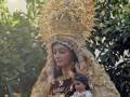 Imagen de una Virgen con un niño en sus brazos, rodeada de velas y adornada con una gran corona de flores. La escena se desarrolla en un entorno natural con árboles al fondo.