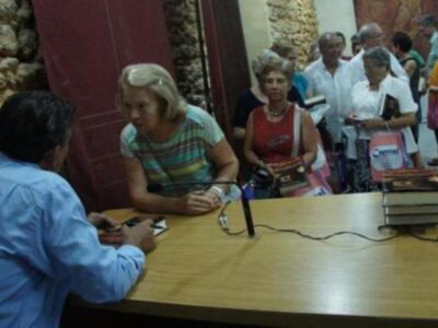 Entrevista en un evento cultural con libros y audiencia en el fondo.