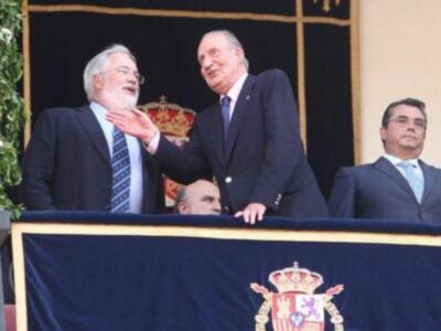 Dos hombres en trajes de negocios saludándose desde un balcón con el escudo español.