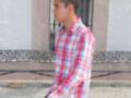 Un joven camina por una calle con un fondo de edificios y una cerca. Wearing a red and white checkered shirt, he has short hair and is looking to the side.