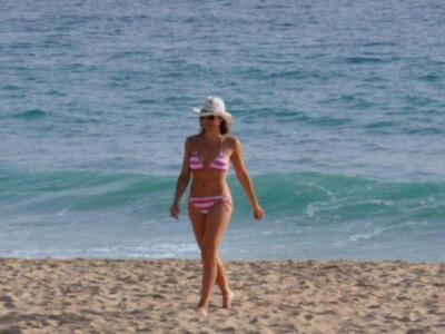 Una mujer en bikini y sombrero blanco camina por la playa, con olas azules en el fondo.