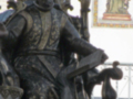 Estatua de bronce de un sacerdote con libro abierto, vestido con traje ornamental, en un entorno urbano con mosaico en el fondo.