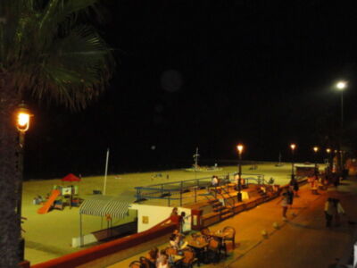 Playa nocturna con sombrillas, palmeras y gente disfrutando.