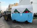 Un tractor verde con un toldo blanco y azul a cuadros lleva una mesa cubierta con mantel azul. La imagen muestra un entorno urbano con edificios y una calle adoquinada.