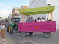 Un tractor verde con una carreta rosa y un toldo verde está en movimiento por una calle. La carreta transporta a varias personas, incluyendo un niño y adultos. Al fondo, se pueden ver edificios y una persona caminando en la calle.