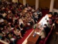 Una sala de conferencias con asistentes sentados en filas, un hombre hablando frente a una mesa con botellas y cuadernos.