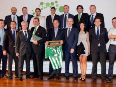 Un grupo de personas posa para una foto en un evento formal, con una bandera verde y blanca en el centro. La escena sugiere una celebración o reconocimiento.