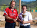 Dos hombres posan con un trofeo en una zona deportiva, rodeados de vegetación y montañas.